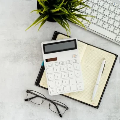 Calculette blanche, cahier ouvert, stylo, lunettes et plante sur un bureau clair.