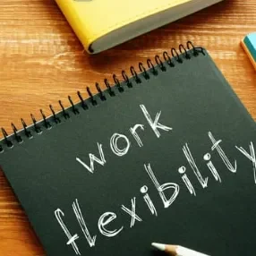 Texte noir sur fond bois : "work flexibility" écrit à la craie. Carnet et crayons en arrière-plan.